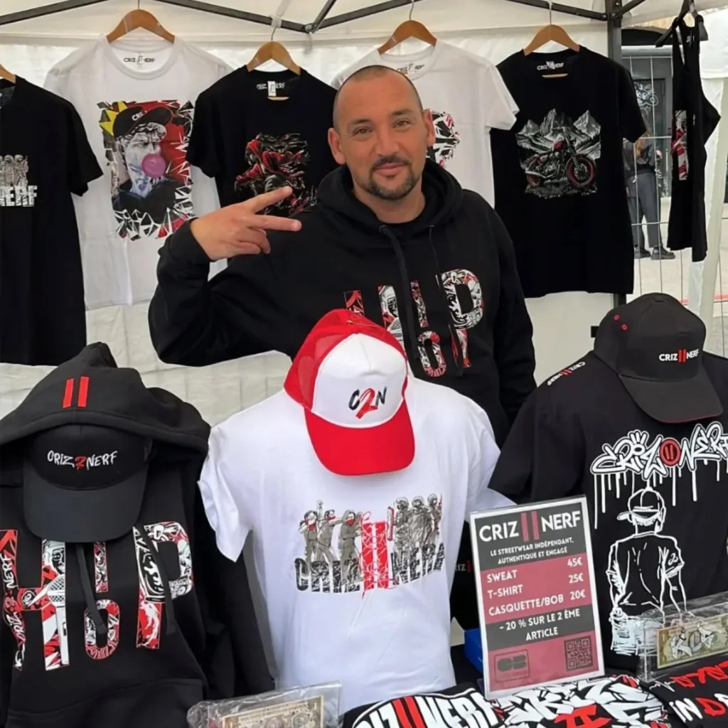 Christophe sur le stand Criz2nerf, entouré de t-shirts et casquettes